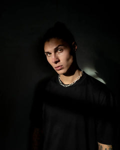 A man wearing a big silver chain necklace with large rectangular links, styled with a black shirt, standing in a dimly lit setting.