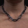 Close-up of a large chain necklace with rectangular black links worn around a person's neck over a black shirt against a dark background.