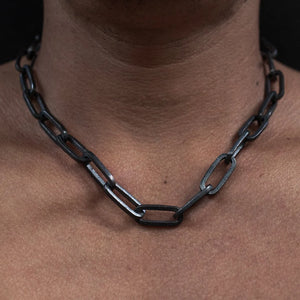 Close-up of a large chain necklace with rectangular black links worn around a person's neck over a black shirt against a dark background.