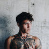 Man with tattoos wearing a Gopa large chain necklace featuring a circular centerpiece, standing against a textured concrete wall.