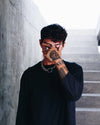 Man wearing a Gopa thick chain necklace with a circular ring, covering his face with a tattooed hand, standing in front of concrete stairs.