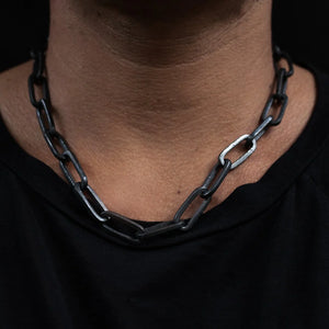 Close-up of a black chunky chain necklace with large rectangular links resting on a person's bare neck against a dark background.