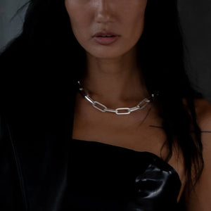 Woman wearing a large chain necklace with rectangular silver links, styled with a black strapless top and dark hair.