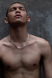 A shirtless man wearing a men's chunky chain necklace with large silver rectangular links, standing against a dark textured background.