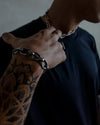 A man wearing a short chunky chain necklace and a matching silver bracelet, shown with a tattooed arm and a black shirt.