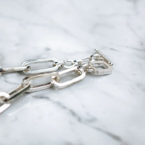 Close-up of a silver chunky chain necklace with rectangular links and a toggle clasp, placed on a marble surface.