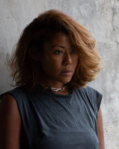 A woman wearing a sterling silver large chain necklace with rectangular links, paired with a grey sleeveless top against a concrete wall.