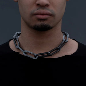 Close-up of a man wearing an XL black chain necklace with large rectangular links, paired with a black shirt against a dark background.